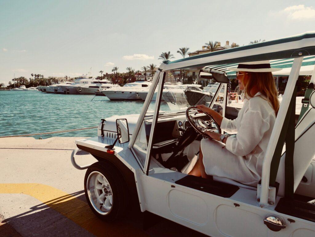 Woman in white dress driving a classic car by the marina with luxury yachts, ideal for family holidays in Sotogrande
