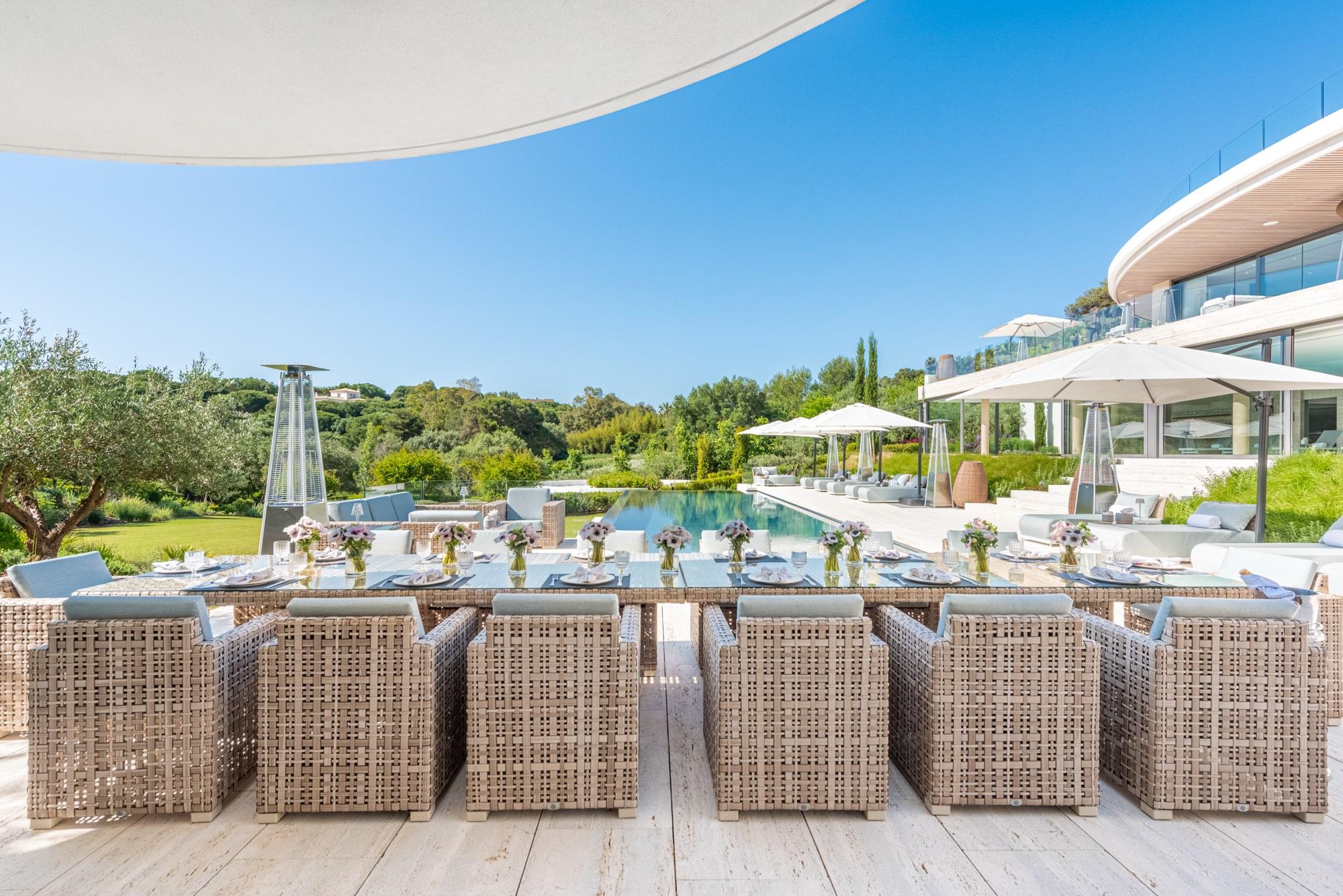 Elegant outdoor dining area with wicker chairs, a long table, and an infinity pool in a luxury Sotogrande property, surrounded by lush greenery.