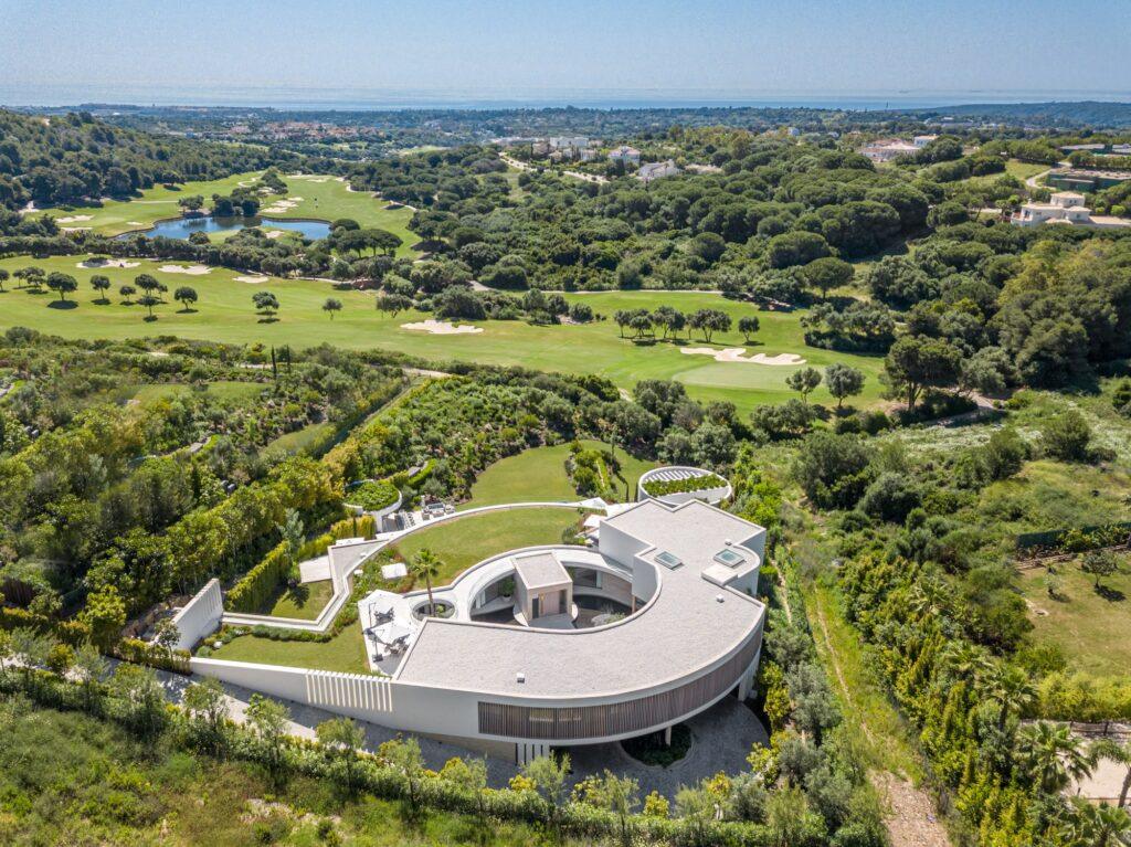 Aerial view of a modern circular building surrounded by lush greenery and golf courses at La Reserva Club.