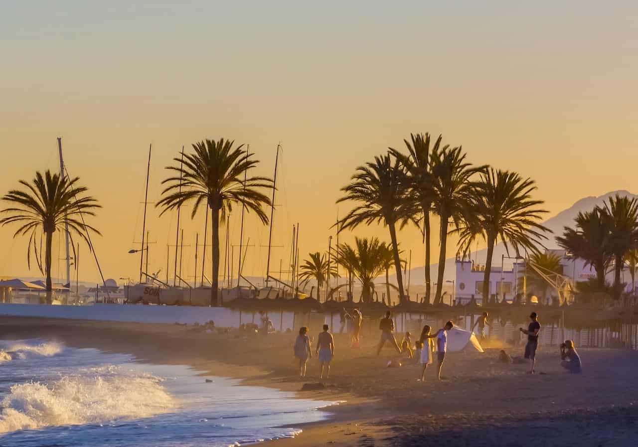 People enjoying a beach sunset in Sotogrande, Spain with palm trees and sailboats in the background, capturing the relaxed Sotogrande lifestyle.