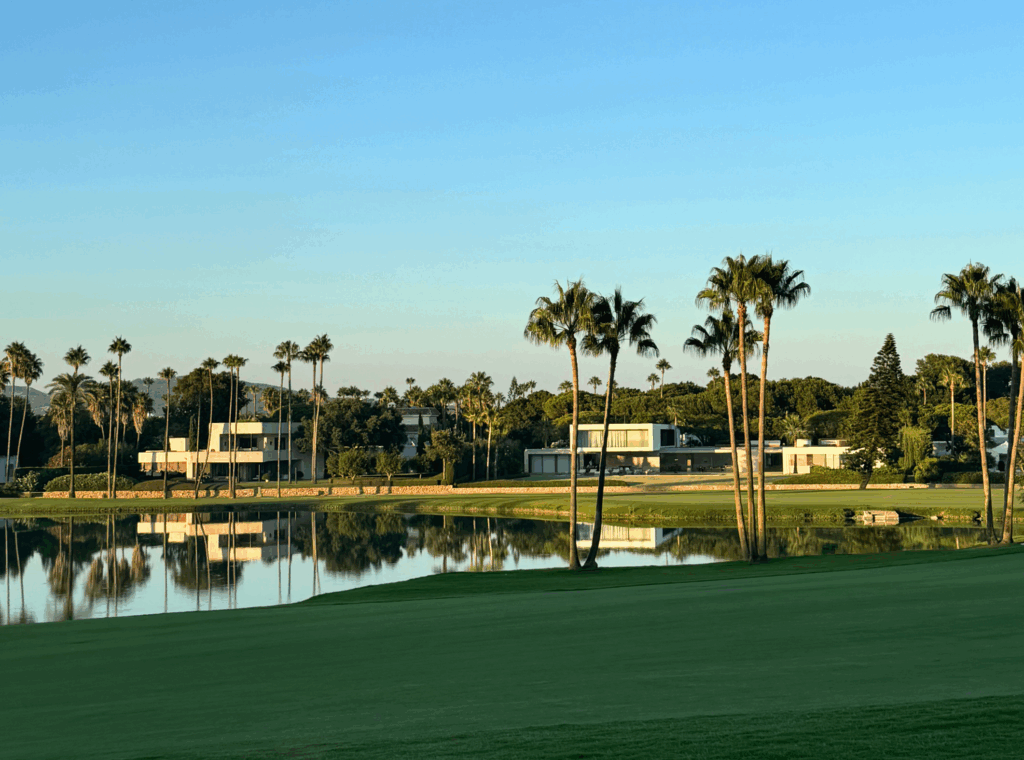 Modern villas along a calm water feature on a golf course in Sotogrande, surrounded by palm trees and lush greenery at sunset.